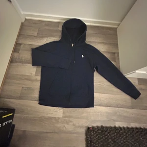 Mörkblå hoodie från Polo Ralph Lauren - Säljer en snygg mörkblå hoodie från Polo Ralph Lauren med dragkedja och huva. Den har en liten vit logotyp på bröstet och praktiska fickor framtill. Perfekt för en avslappnad stil.