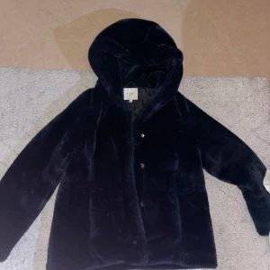 Svart pälsjacka från Cellbes - Mysig svart pälsjacka från Cellbes med huva och knappar framtill. Perfekt för kyliga dagar när du vill hålla dig varm och stilren.