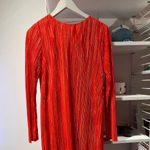 Orange  plisserad klänning från H&M - Snygg orange plisserad klänning från H&M med långa ärmar och en knappdetalj i nacken. Perfekt för en färgstark look. Klänningen har en elegant och flytande design. Färgen kommer inte till sin rätt på bilderna. På bilderna ser klänningen randig eller flammig ut. Det är den inte. 