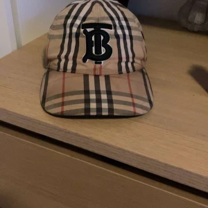 Rutig keps från Burberry - Snygg rutig keps från Burberry med klassiskt mönster i beige, svart och rött. Kepsen har en broderad logotyp framtill och justerbar passform. kan bytas mot en gucci keps 