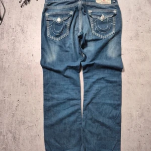 Blå jeans från True Religion - Snygga blå jeans från True Religion med kontrasterande vita sömmar och klassiska femficksdesign. Byxorna har en knappdetalj på bakfickorna och en vintageinspirerad etikett. Perfekta för en avslappnad stil.