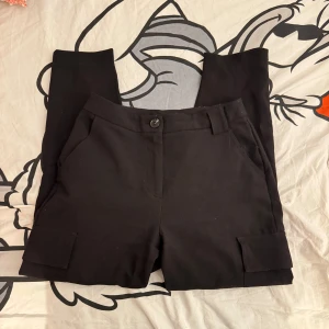 Svarta kostymbyxor med fickdetaljer - Snygga svarta kostym liknande byxor med extra fickor och knappdetaljer på änderna. I storlek 36. 