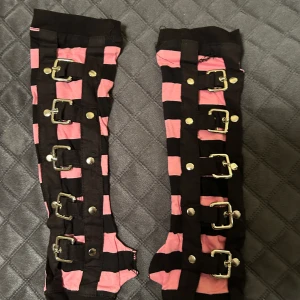 Svart och rosa randiga handskar med spännen - Unika byxor med svart och rosa randigt mönster. De har flera dekorativa spännen och knappar längs benen, vilket ger en edgy look. Perfekta för dig som vill sticka ut!