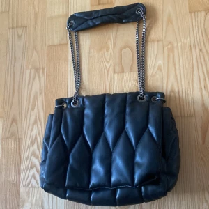 Svart quiltad axelväska från Zara - Snygg svart quiltad axelväska från Zara med kedjedetaljer på axelremmen. Väskan har en stilren design med tryckknappar för stängning och en mjuk yta. Perfekt för att ge en elegant touch till din outfit.