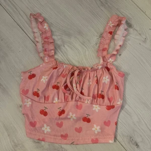 Rosa ribbad linnetopp med körsbärsmönster - Söt rosa ribbad linnetopp med körsbär och hjärtan. Toppen har volangdetaljer på axelbanden och en knytning framtill. Perfekt för sommarens varma dagar!