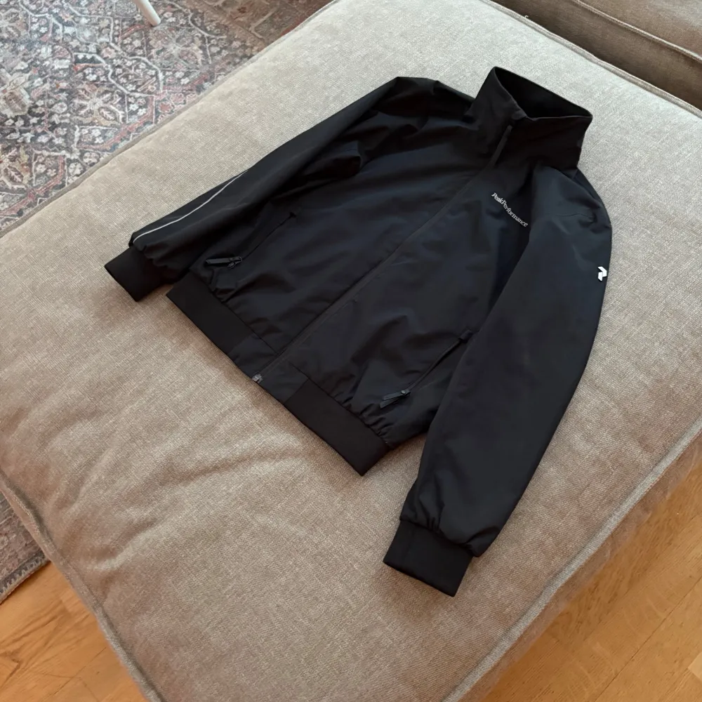 Snygg svart vindjacka från Peak Performance med dragkedja och hög krage. Jackan har långa ärmar med ribbade muddar och två praktiska fickor med dragkedja. Perfekt för blåsiga dagar.. Takit.