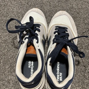 New Balance 997H sneakers - Snygga New Balance 997H sneakers i vitt och marinblått med bruna detaljer. Skorna har en klassisk design med snörning och en bekväm sula. Perfekta för en stilren och sportig look.