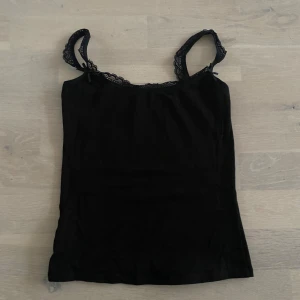 Svart linne med spetsdetaljer - Snyggt svart linne med spetsdetaljer vid axelbanden. Perfekt för en stilren look. Axelbandslös design som ger en elegant touch. Aldrig använd 