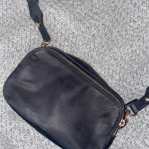 Svart axelväska med dragkedja - Snygg svart axelväska med justerbar rem och två dragkedjor i guld. Perfekt för att hålla dina saker organiserade och lättillgängliga. Insidan är ljus och rymlig, vilket gör den praktisk för vardagsbruk.