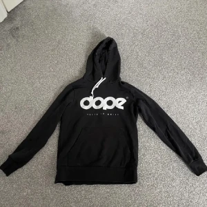 Svart hoodie från Dope - Säljer en svart hoodie från Dope med vit logga på bröstet. Den har en stor ficka framtill och justerbar huva med vita snören. Inte använt så mycket och vill sälja då jag bytt stil! (Lånad bild) Storlek S/XS vet inte exakt då jag klippt bort lappen
