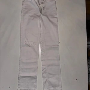 Vita jeansbyxor från NA-KD - Snygga vita jeansbyxor från NA-KD med en trendig rippad detalj på ena benet. Byxorna har en klassisk femficksdesign och stängs med dragkedja och knapp. Perfekta för en stilren look.