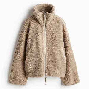 Ny Beige teddyjacka i storlek S från H&M - Ny och oanvänd med etiketter kvar. En beige tjock och varm teddyjacka i mjuk pile med kantband i ytbehandlad kvalitet. Jackan har lös passform, ståkrage, tvåvägsdragkedja fram och kängurufickor. Fodrad och lätt vadderad.  Storlek: S (något oversized) Färg: Beige, Årets trendfärg för 2025 är Mocha mousse