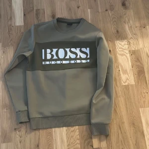 Hugo boss Sweatshirt - Säljer en stilren olivgrön sweatshirt från Hugo Boss med stort vitt logotryck på bröstet. Tröjan har långa ärmar och ribbade muddar vid ärmslut och nederkant. Perfekt för en avslappnad stil. Mycket bra skick!