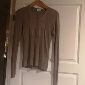 Beige ribbad tröja från Tiger of Sweden - Säljer en stilren beige ribbad tröja från Tiger of Sweden. Tröjan har långa ärmar och knappar framtill som ger en klassisk look. Perfekt för en avslappnad men ändå elegant stil.