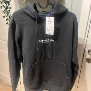 HELT NY!  Mörkgrå hoodie från Jack & Jones  - Säljer en snygg mörkgrå hoodie från Jack & Jones med texten 'Originals Studio' på bröstet. Den har en klassisk känguruficka och justerbar dragsko i huvan. Perfekt för en avslappnad stil.