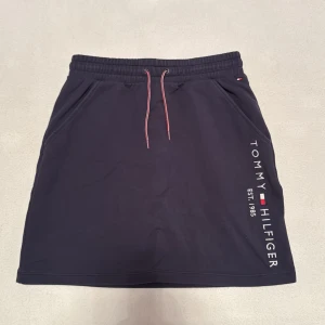 Mörkblå kjol från Tommy Hilfiger - Inte så använd kjol med fickor. Snygg mörkblå kjol från Tommy Hilfiger med elastisk midja och dragsko. Kjolen har en stilren design med logotypen tryckt längs sidan. Perfekt för en avslappnad och sportig look.