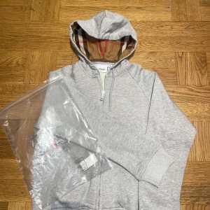 Grå hoodie från Burberry - Säljer en stilren grå hoodie från Burberry med klassiskt rutigt foder i huvan. Tröjan har dragkedja framtill och ribbade muddar vid ärmslut och nederkant. Perfekt för en avslappnad och trendig look.