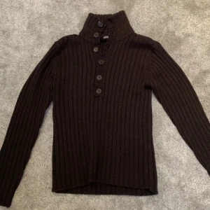 Brun ribbad tröja från H&M - Säljer en stilren svart ribbad tröja från H&M med hög krage och knappar framtill. Perfekt för kyligare dagar med sin långärmade design.