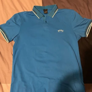 pikétröja från Boss - Snygg blå pikétröja från Boss med klassisk krage och knappar. Tröjan har vita och mörkblå ränder på kragen och ärmsluten. Då den inte kommer till användning längre.