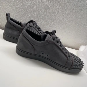 Grå sneakers med nitar från Christian Louboutin - Snygga grå sneakers från Christian Louboutin med nitar på tån och den ikoniska röda sulan. Skorna har snörning och är tillverkade i skinn.