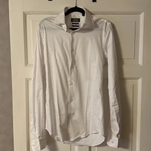 Vit skjorta från Dressmann - Snygg vit skjorta från Dressmann i slim fit med extra lång passform i ärmar. Tillverkad i premium bomullsstretch för en bekväm känsla. Perfekt för en stilren look. Perfekt att ha under en kavaj eller cardigan. Använd en gång.