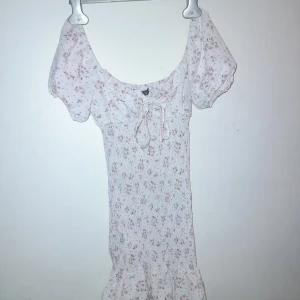 Blommig klänning från Shein - Söt blommig klänning från Shein i vitt med rosa och gröna blommor. Klänningen har korta puffärmar och en volangdetalj vid fållen. Den har en knytning framtill och en smickrande passform.