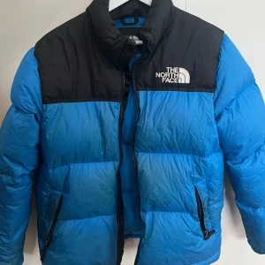Blå och svart dunjacka från The North Face - Säljer en blå och svart dunjacka från The North Face. Jackan har en hög krage och dragkedja framtill. Perfekt för kyliga dagar med sin pufferdesign och varma fyllning.