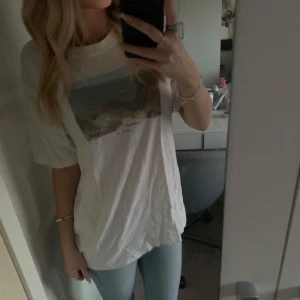 Vit t-shirt med tryck från Gina Tricot - Säljer en vit t-shirt från Gina Tricot med ett tryck av ett kustlandskap i blå och gröna toner. T-shirten har en normal passform och korta ärmar. Perfekt för en avslappnad stil. Den sitter baggy på mig då den är i M (jag har storlek xs/s i topp). Knappt använd, är i nyskick🙃