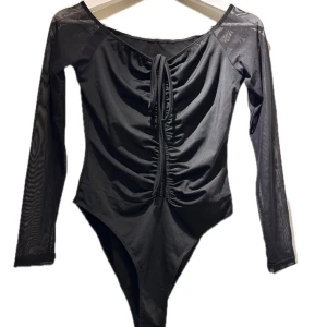 Svart långärmad bodysuit - Snygg svart bodysuit med långa meshärmar och knytning framtill. Perfekt för en kväll ute eller en stilren vardagslook. Den har en åtsittande passform och en elegant design.