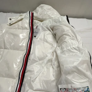 Vit pufferjacka från Moncler - Snygg vit pufferjacka från Moncler med glansig finish och ikonisk logga på bröstet. Jackan har en praktisk huva och dragkedja framtill med röd och svart randdetalj. Perfekt för kyliga dagar.