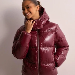 Vinröd pufferjacka nelly  - Snygg vinröd pufferjacka med dragkedja och hög krage. Perfekt för kyliga dagar med sin vadderade design som håller dig varm och bekväm. Jackan har en glansig finish som ger en modern touch. Den är endast använd en gång 