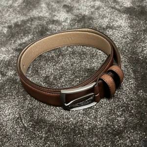 Brunt skärp med silverfärgat spänne - Säljer ett snyggt brunt skärp i skinn och mocka med ett stilrent silverfärgat spänne. Perfekt för att ge en klassisk touch till vilken outfit som helst. Skärpet har flera hål för justerbar passform och är i nyskick.