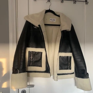 Svart och beige jacka från NA-KD - Säljer en supercool svart och beige jacka från NA-KD. Den är i storlek 40 och har en snygg design med dragkedja och bältesdetaljer vid ärmsluten. Jackan är fodrad med mjukt beige material som ger en skön och varm känsla. Perfekt för höst och vinter!