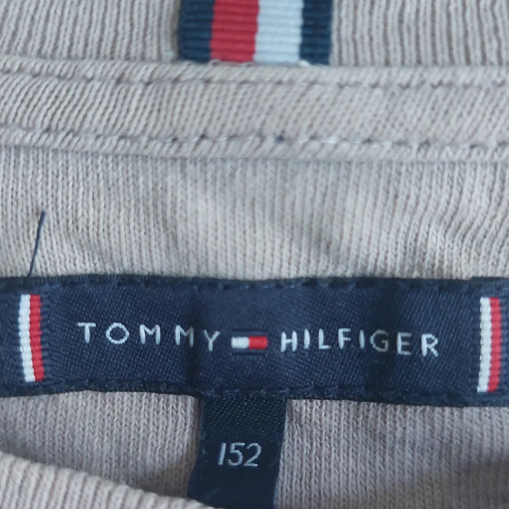 Säljer en snygg beige t-shirt från Tommy Hilfiger. Den har en klassisk rund hals och korta ärmar. På framsidan finns en broderad Tommy-logga i rött, vitt och blått. Perfekt för en avslappnad stil! OPS har två små hål på framsida  och ett på baksidan.. T-shirts.