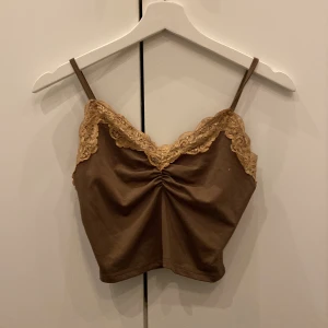 Brun linne med spetsdetaljer - Säljer en superfin brun linne med smala axelband och vacker spetsdetalj längs urringningen. Toppen har en snygg rynkning framtill som ger en extra touch. Perfekt för sommardagar eller som ett lager under en kavaj. Materialet känns mjukt och bekvämt mot huden.