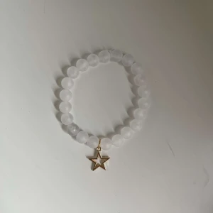 Vitt armband med stjärnhänge - Säljer ett superfint armband med vita pärlor och ett gulligt stjärnhänge i guld. Perfekt för att ge en extra touch till din outfit! Passar både till vardags och fest. ✨