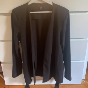 Svart kavaj från Amisu - Säljer en stilren svart kavaj från Amisu. Den har långa ärmar och ett bälte i midjan för en snygg passform. Perfekt för både vardag och fest!