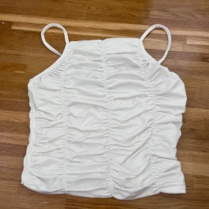 Vit ärmlös topp med rynkdetaljer - Säljer en superfin vit topp med smala axelband och rynkdetaljer både fram och bak. Toppen är ärmlös och har en tight passform som är perfekt för sommaren. Materialet känns mjukt och bekvämt, perfekt för varma dagar!