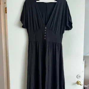 Svart klänning med knappar - Säljer en elegant svart klänning med v-ringning och knappar framtill. Klänningen har korta ärmar och en bekväm passform som passar perfekt för både vardag och fest. Den är gjord av ett mjukt och följsamt material.