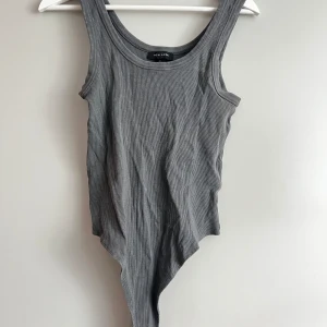 Grå ribbad bodysuit från New Look - Säljer en snygg grå ribbad bodysuit från New Look. Den är ärmlös och har en tight passform, perfekt för att styla med jeans eller kjol. Materialet är mjukt och stretchigt, vilket gör den bekväm att bära hela dagen.