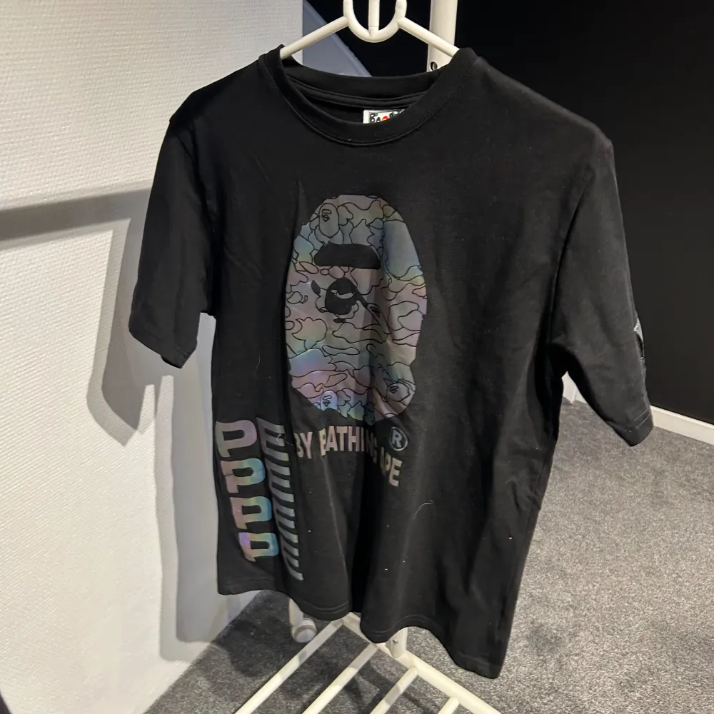Säljer en svart t-shirt från A Bathing Ape med deras ikoniska logga i ett coolt, färgglatt mönster på framsidan. T-shirten har korta ärmar och texten 'BY BATHING APE' under loggan. På sidan finns det flerfärgad text 'APE' som ger en extra touch. Perfekt för streetwear-stilen! Är storlek L men upplevs mindre. T-paidat.