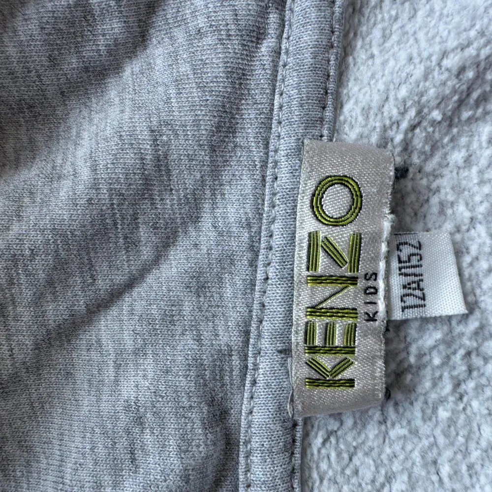 Säljer en supercool grå hoodie från Kenzo Kids. Den har ett stort tryck på framsidan med Kenzo-loggan i rött och en blå design. Perfekt för att hålla sig varm och stilren. Den är i bra skick och redo för en ny ägare!. Tröjor & Koftor.