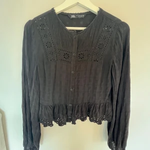 Svart blus från Zara - Säljer en elegant svart blus från Zara. Den har långa ärmar och fina spetsdetaljer både vid halsen och nederkanten. Blusen är knäppt framtill och har en lätt volang vid midjan. Perfekt för både vardag och fest! Säljer för att den är för liten