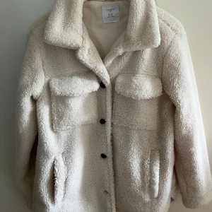 Vit teddyjacka från Lindex - Mysig och fluffig vit teddyjacka från Lindex. Perfekt för kyliga dagar med sin mjuka och varma känsla. Jackan har knappar framtill och praktiska fickor. En stilren och bekväm jacka för alla tillfällen!