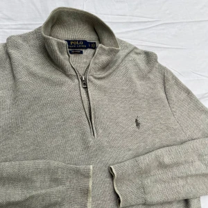 Polo Ralph Lauren half-zip grå - Sparsamt använd och väl omhändertagen. OBS! Den vita bakgrunden får plagget att se mörk och smutsig ut! Stickad half-zip från Polo Ralph Lauren. Tillverkad i bomull i en texturerad stickning. Butikpris: 2 199kr