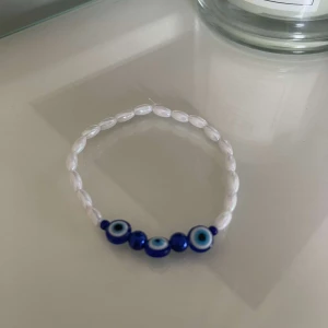 Armband med blå ögonpärlor - Säljer ett snyggt armband med vita pärlor och blå ögonpärlor. Perfekt för att ge en touch av mystik och stil till din outfit. Armbandet är elastiskt och passar de flesta handleder.