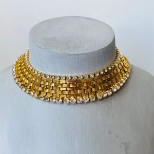 Guldhalsband med glittriga stenar - Säljer ett superglammigt guldhalsband med en bred design och massor av glittriga stenar. Halsbandet har en flätad struktur som ger det en lyxig känsla och är perfekt för festliga tillfällen. Det är både elegant och iögonfallande, och kommer definitivt att dra blickarna till sig! Skapat av eftertraktade designern Mimi di N