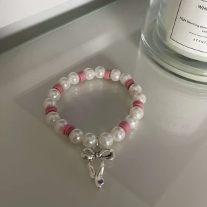 Pärlarmband med rosett - Säljer ett supergulligt armband med vita pärlor och rosa detaljer. Armbandet har en söt liten rosett som hänge, vilket ger det en extra charmig touch. Perfekt för att piffa upp vilken outfit som helst!