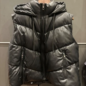 Svart dunväst från Zara (kan gå ner i pris vid snabbaffär) - Säljer en snygg och trendig svart dunväst från Zara. Västen har en cool quiltad design med dragkedja framtill och praktiska fickor med dragkedjor. Den har också en huva som ger extra värme och stil. Perfekt för höst och vinter!