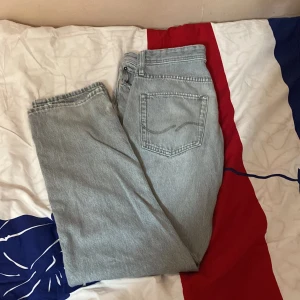 Grå jeans från Jack & Jones (RELAXED/ CHRIS)  - Säljer ett par grå jeans från Jack & Jones. De har en klassisk femficksdesign och en knappgylf. Jeansen är i en ljusgrå nyans och verkar vara i välanvänt skick. Perfekta för en avslappnad vardagslook jeansen har även ett litet hål vid knäppen men de syns inte för man kan ha tröja över!